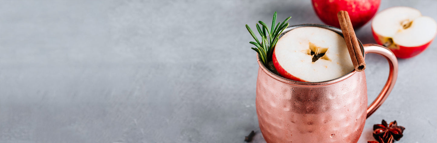 Copper mug with apple cider, garnished with apples and a cinnamon stick, on a gray background.