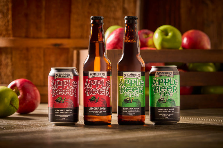 Four bottles and cans of SodaBeers Apple Beer on a wooden surface with apples in the background.