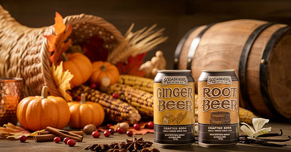 Two cans of Sodastream ginger beer and root beer on a table with pumpkins, corn, and a barrel in the background.