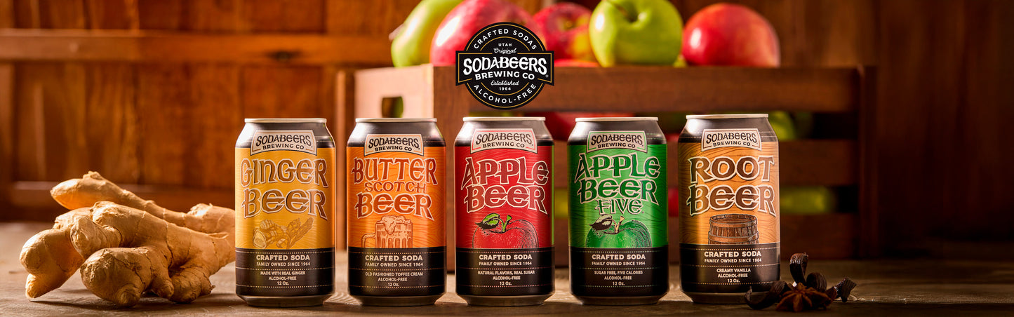 Row of SodaBeers Ginger Beer, Butterbeer, Applebeer, and Root Beer cans with ginger root and apples on a wooden surface.